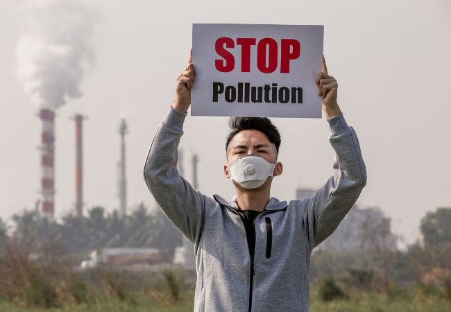 immagine di uomo con mascherina protettiva che alza un cartello con scritto "stop all'inquinamento" davanti ad una fabbrica in funzione