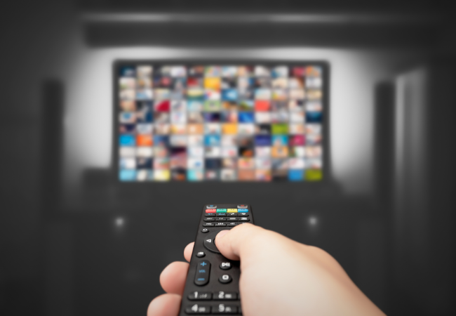 Immagine di un mano che impugna un telecomando con un televisore davanti.