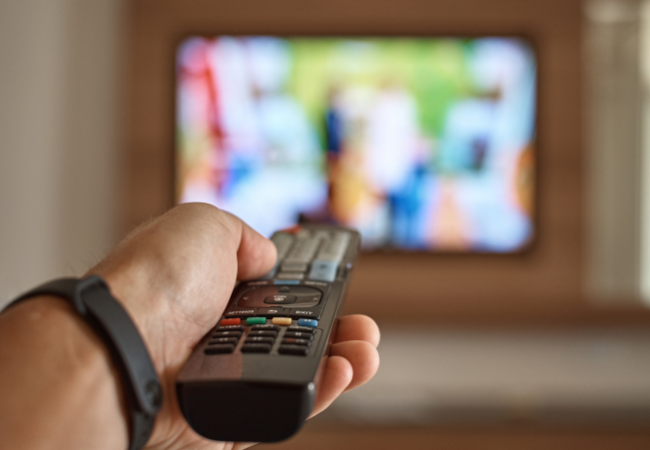 Immagine di un mano che impugna un telecomando con una tv nello sfondo.