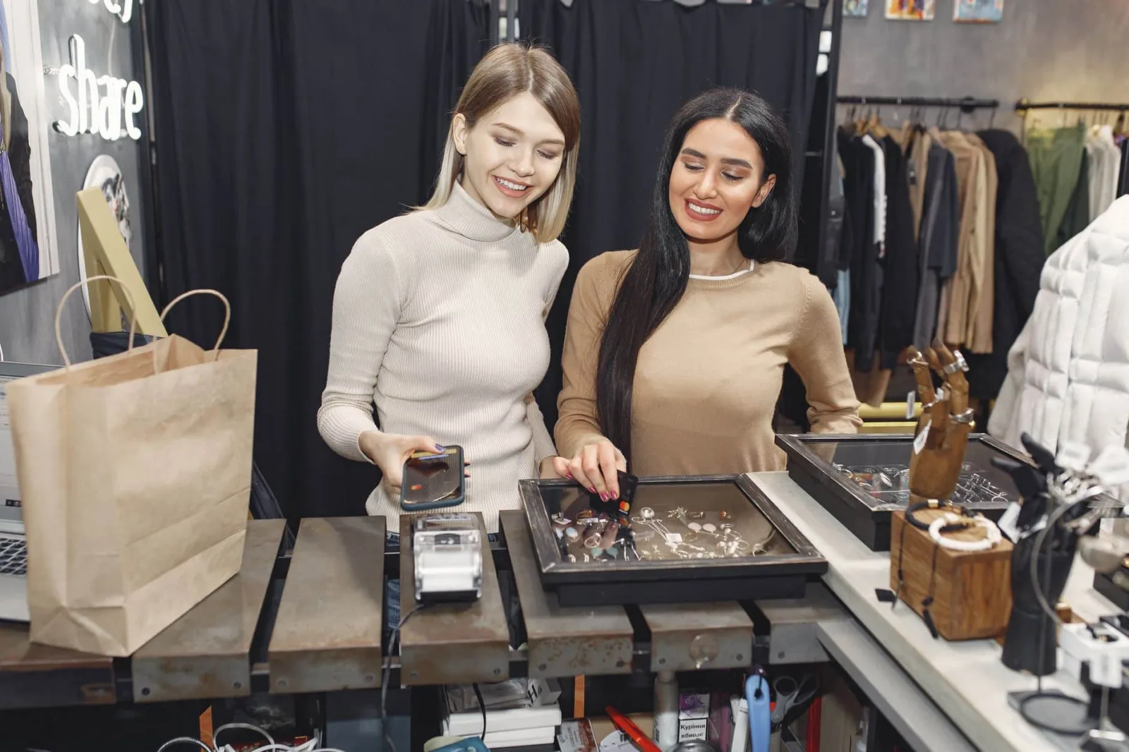 Cheerful casually dressed women paying with smartphone and credit card for purchases while standing near contactless terminal at counter desk