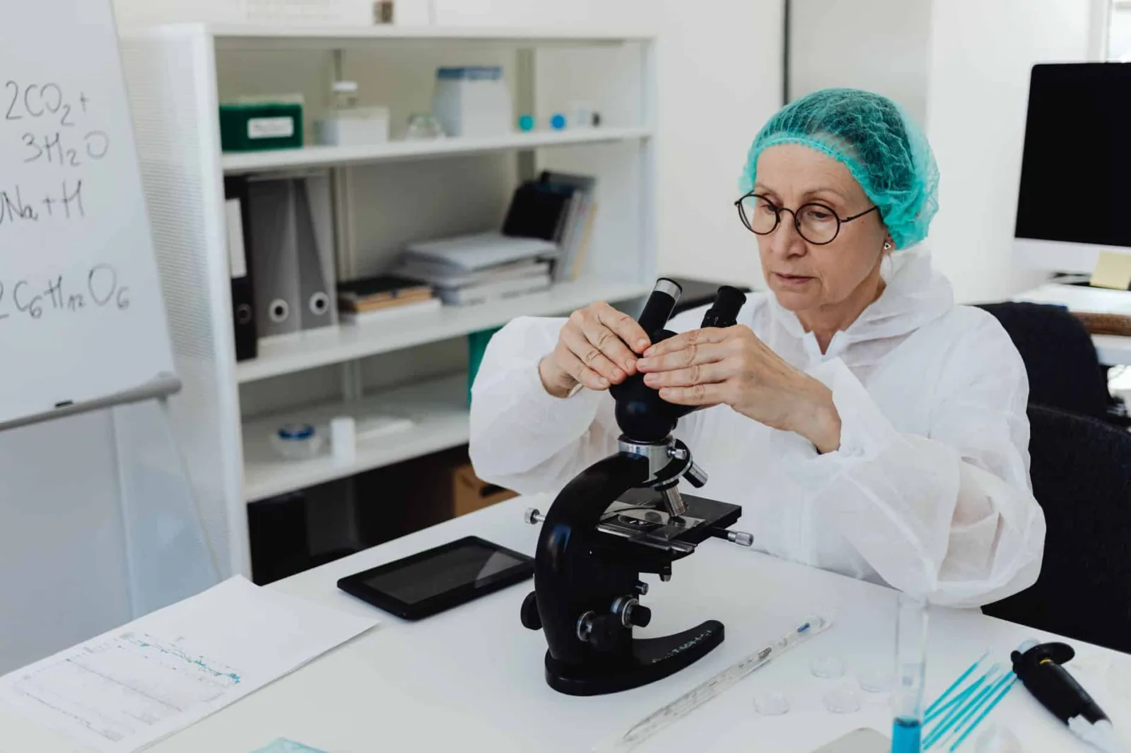 Elderly female scientist in PPE examining samples with a microscope in a modern lab.