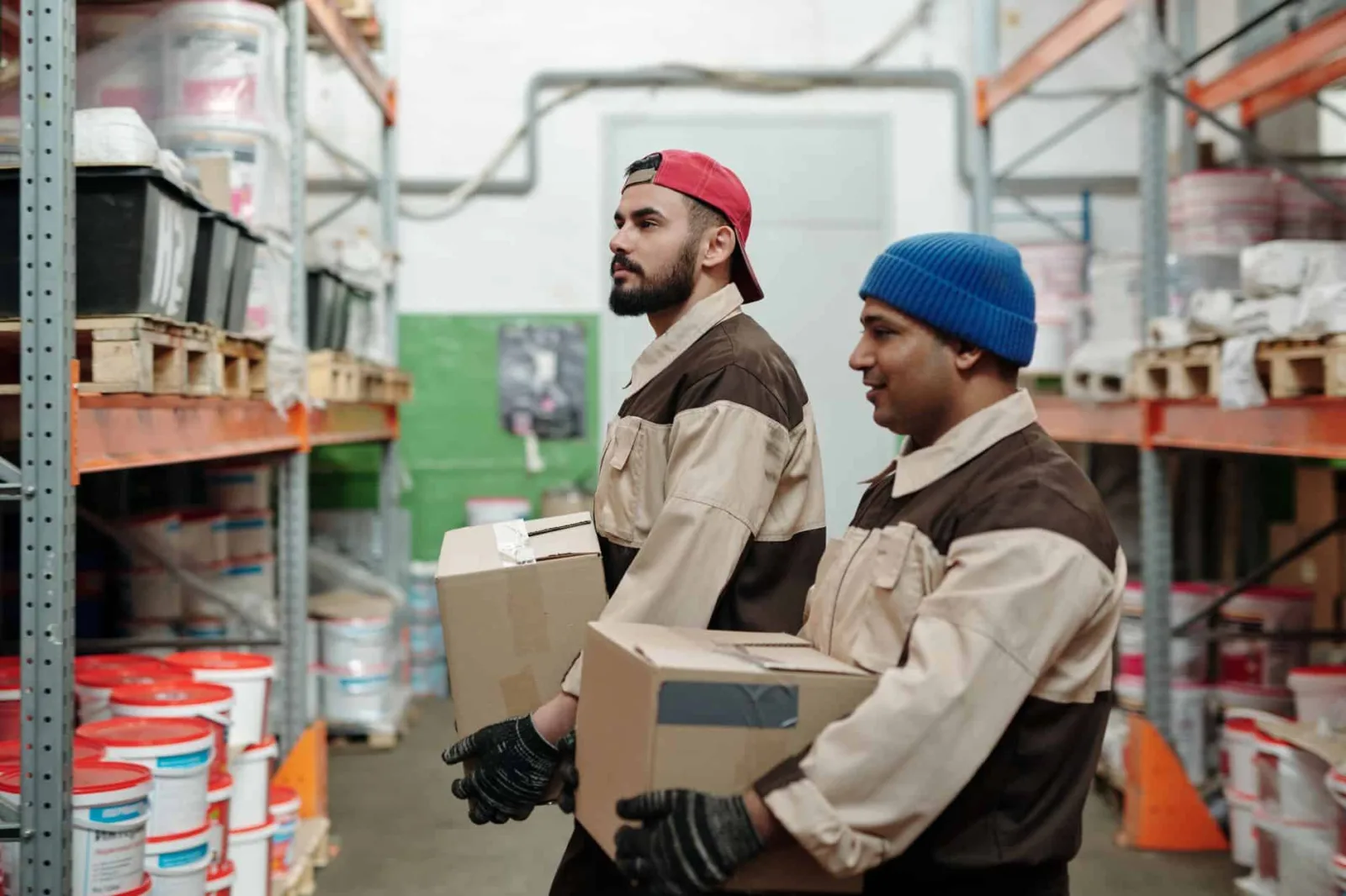 Two workers in a warehouse carrying boxes, organized shelves in the background.