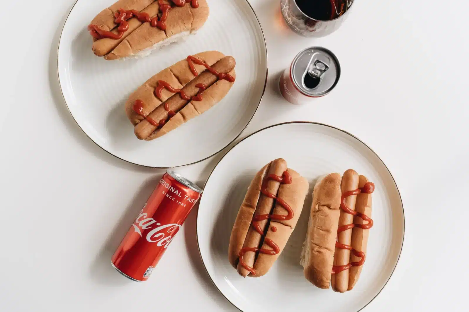 Delicious hot dogs with ketchup paired with Coca Cola cans on plates in a flat lay setting.