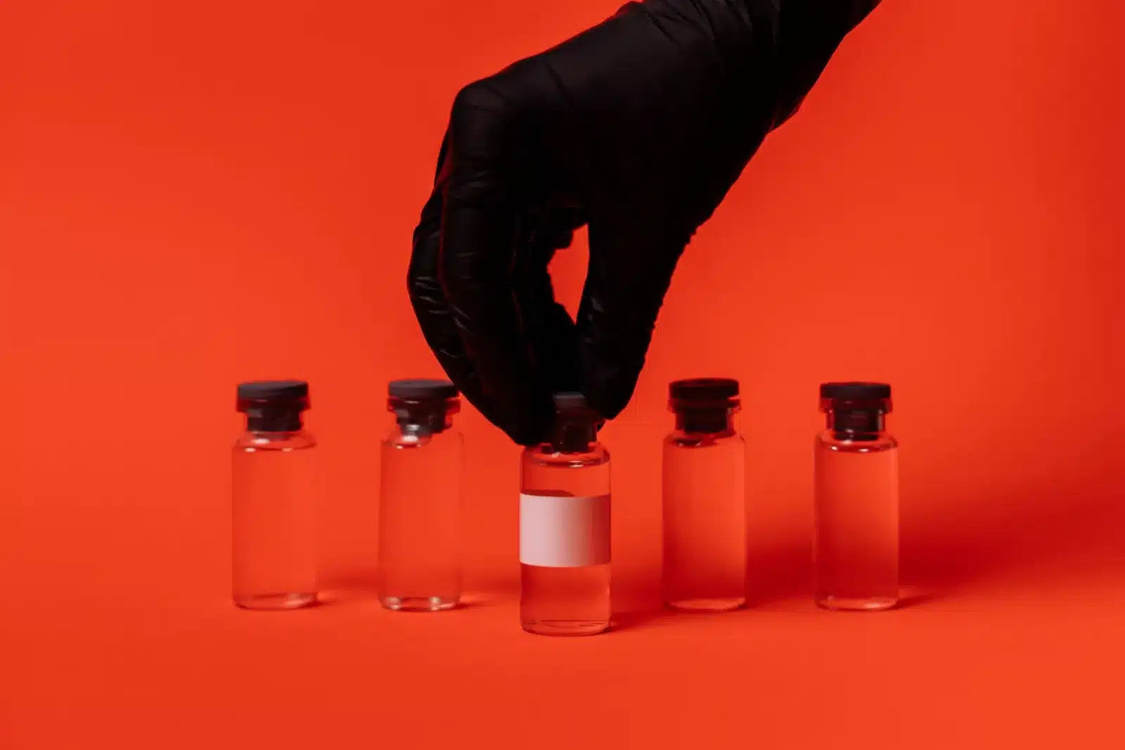 Person wearing black glove grabbing a vaccine vial against a bright red backdrop.